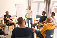 Eine DRK-Mitarbeiterin steht vor einer Pin-Wand und erklärt Sachverhalte den in einem runden Kreis um sie herumsitzenden Gruppe an Männern.
