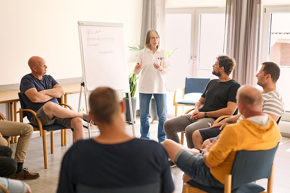 Schulung Eine DRK-Mitarbeiterin steht vor einer Pin-Wand und erklärt Sachverhalte den in einem runden Kreis um sie herumsitzenden Gruppe an Männern.