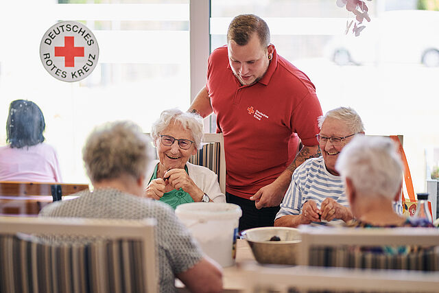 Mehrere Senioren sitzen gemeinsam an einem Tisch und werden durch einen Helfer vom Roten Kreuz bedient