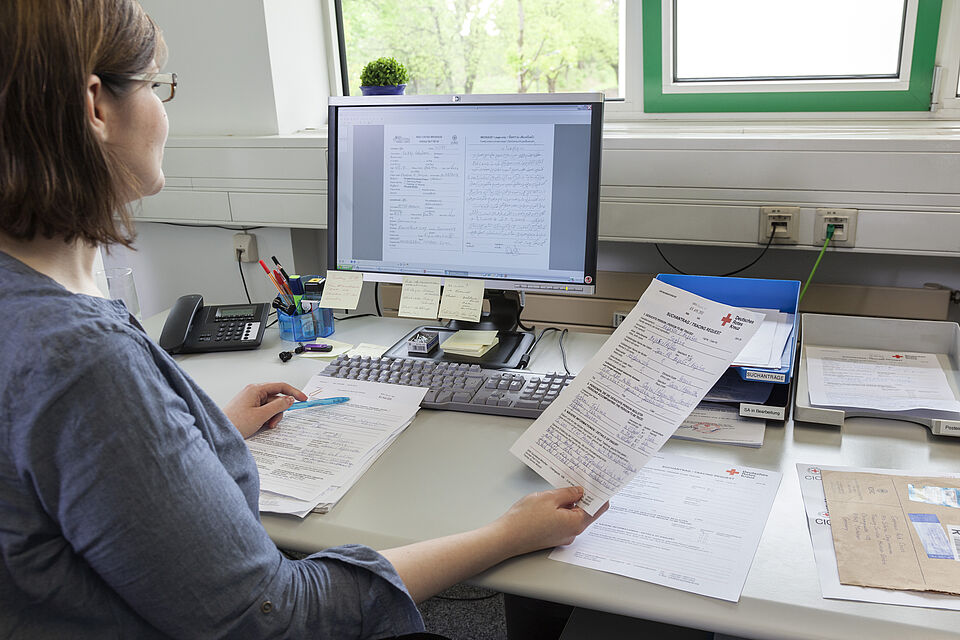 Eine Frau sitzt vor einem aufgeklappten Laptop und hält ein Blattpapier in der Hand wovon sie Inhalte abließt die von ihr in den Computer übertragen werden.