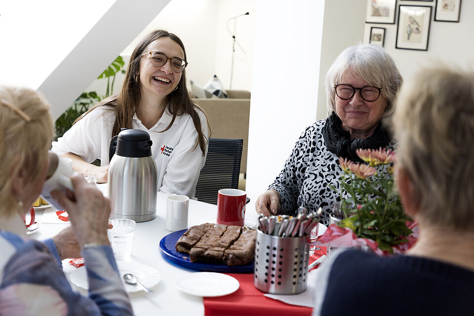 Es sitzen mehrere Frauen an einem gedeckten Tisch. Es stehen mehrere Tassen, eine Kaffeekanne und Kuchen auf dem Tisch. Es wird gelacht. Eine junge Frau, die am Tisch sitzt, trägt ein weißes Polo-Shirt mit DRK Emblem.