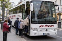 Eine Gruppe von Senioren steigt in einen weißen Reisebus ein. Eine Frau im roten Jacket steht vor dem Bus und notiert dei Anwesenheit der Reiseteilnehmer. Der Busfahrer steht neben der Türe.