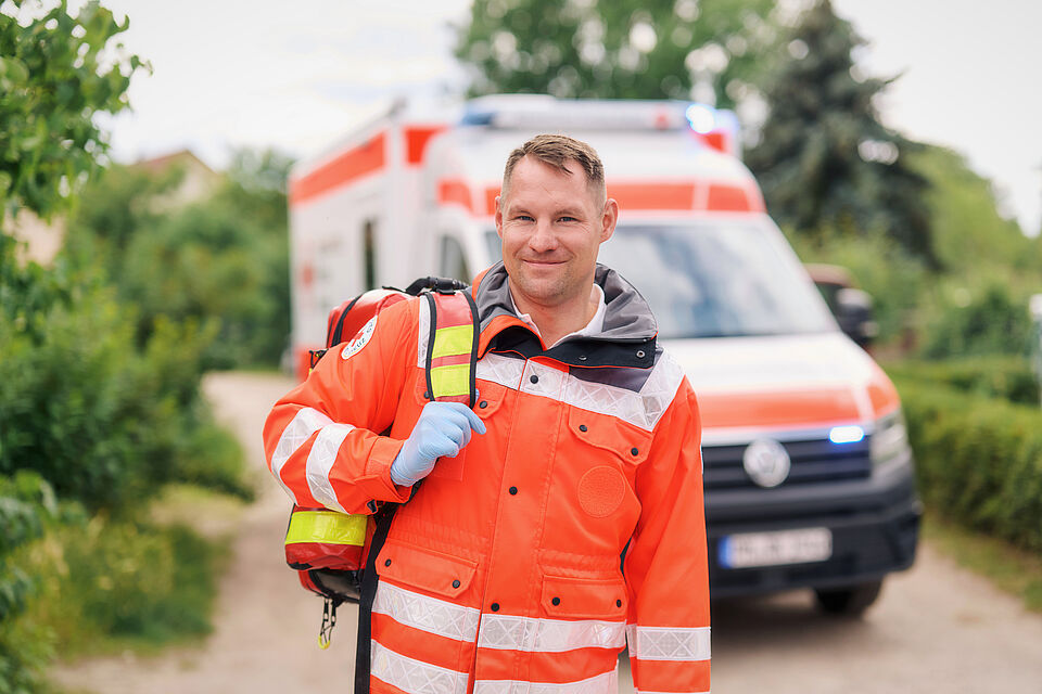 Notfallsanitäter des DRK Ein Notfallsanitäter in oranger Schutzkleidung kommt einem mit geschultereter Notfalltasche entgegen. Im Hintergrund ist ein DRK Rettungswagen zu sehen.